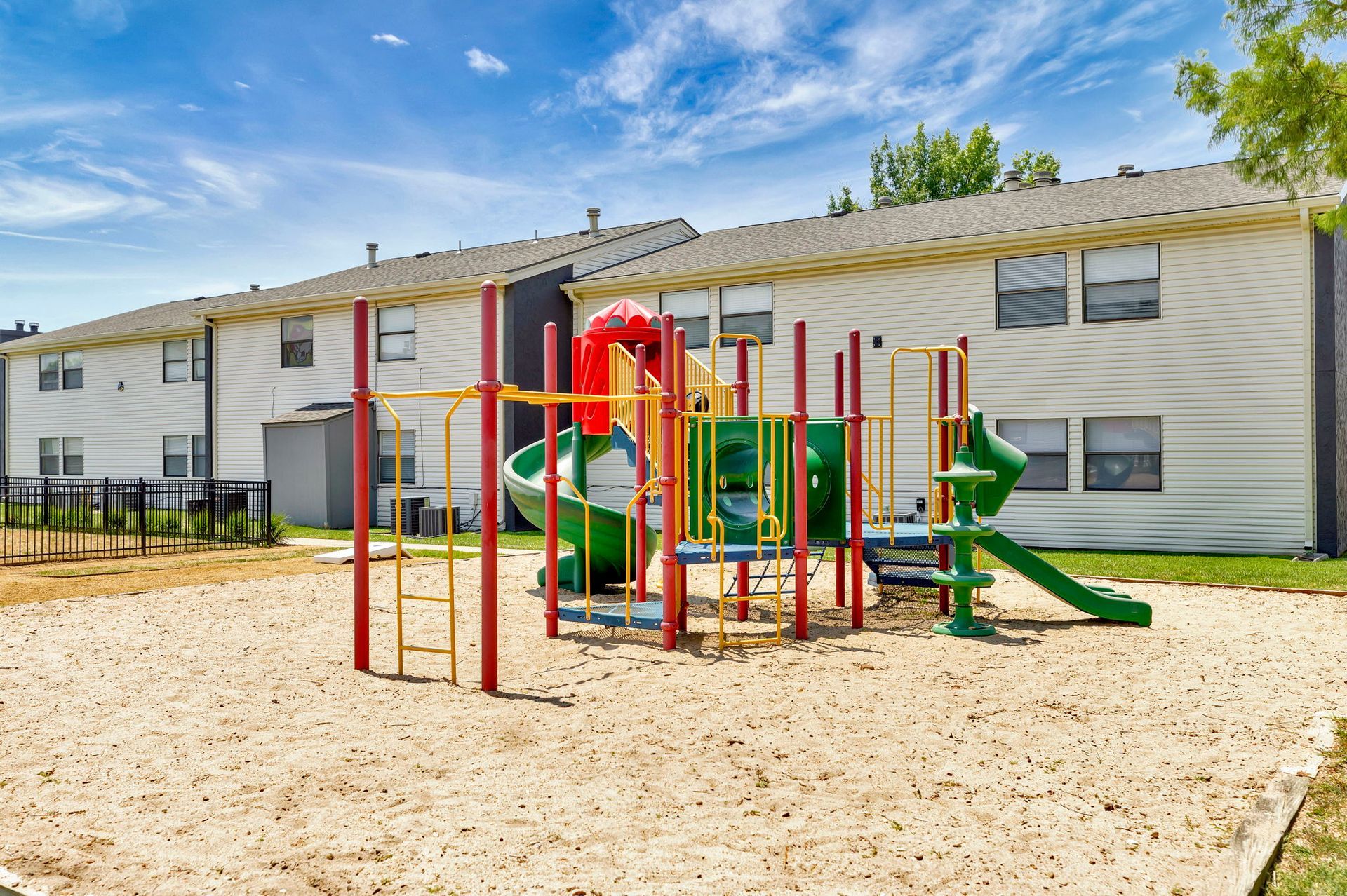 Playground in front of two-story apartment building, with sand ground cover, slides, and climbing structures.