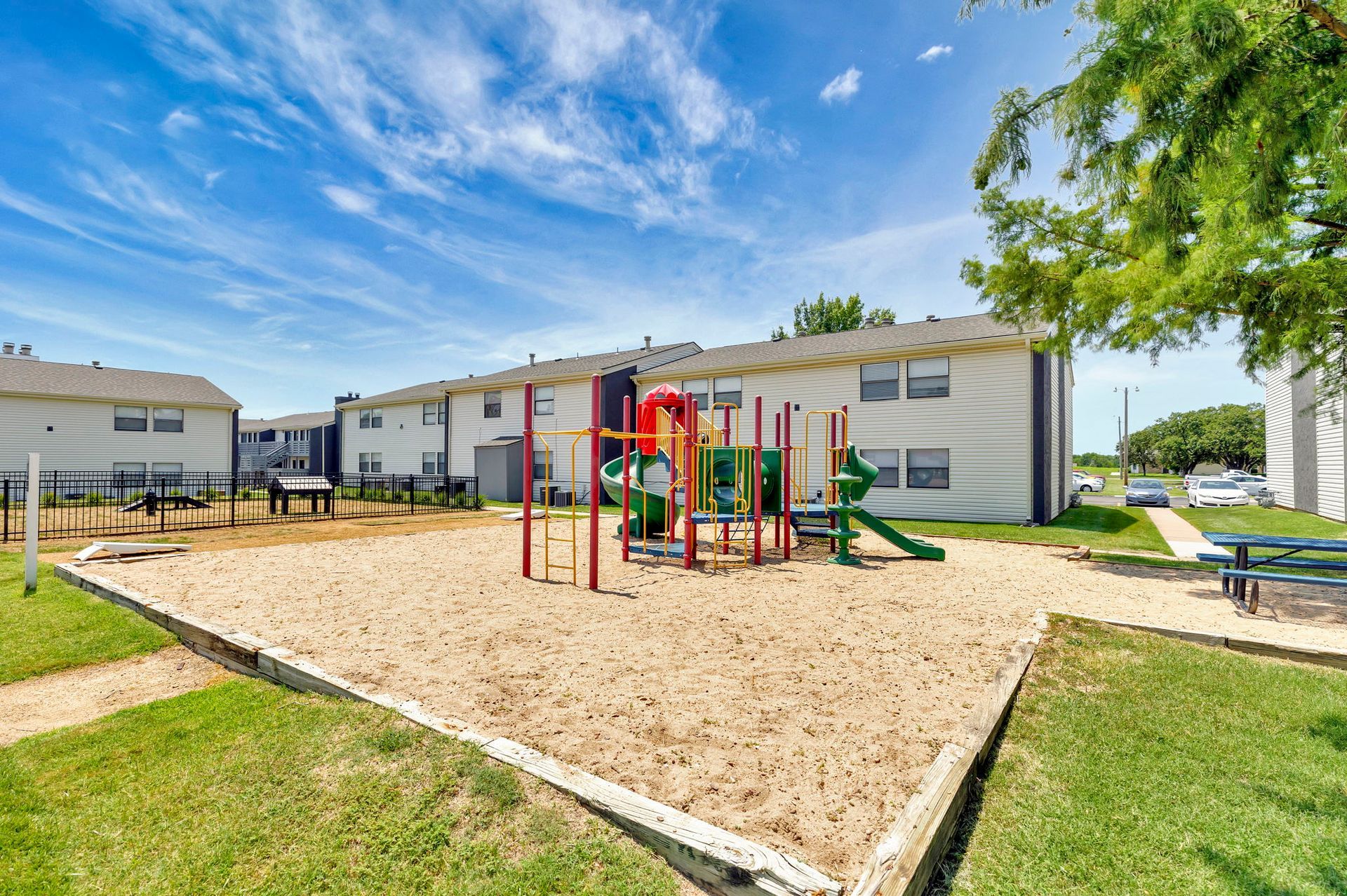 Playground with slide, climbing structures, and sandbox in front of apartment buildings on a sunny day.