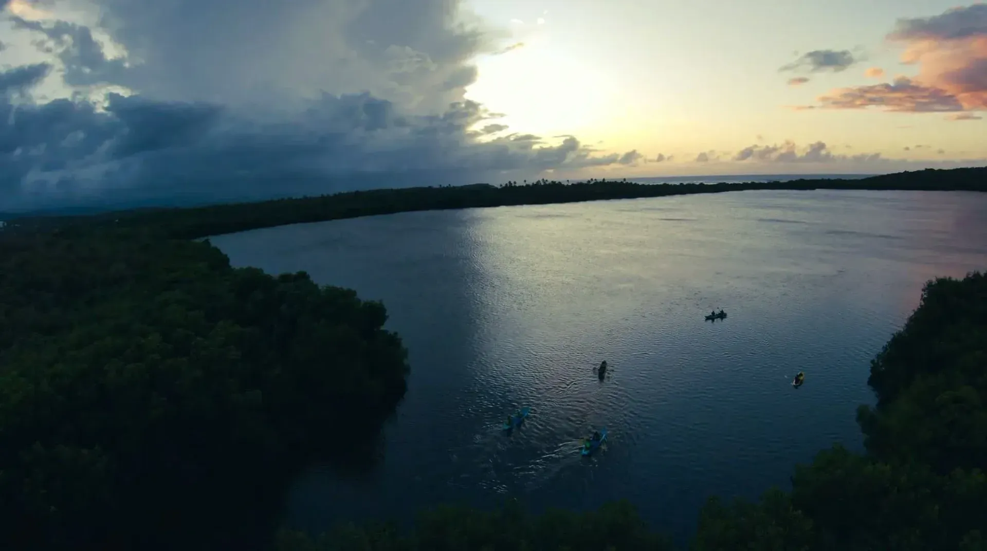 Sunset over a calm lake framed by dark shoreline and clouds, with a small boat near the center