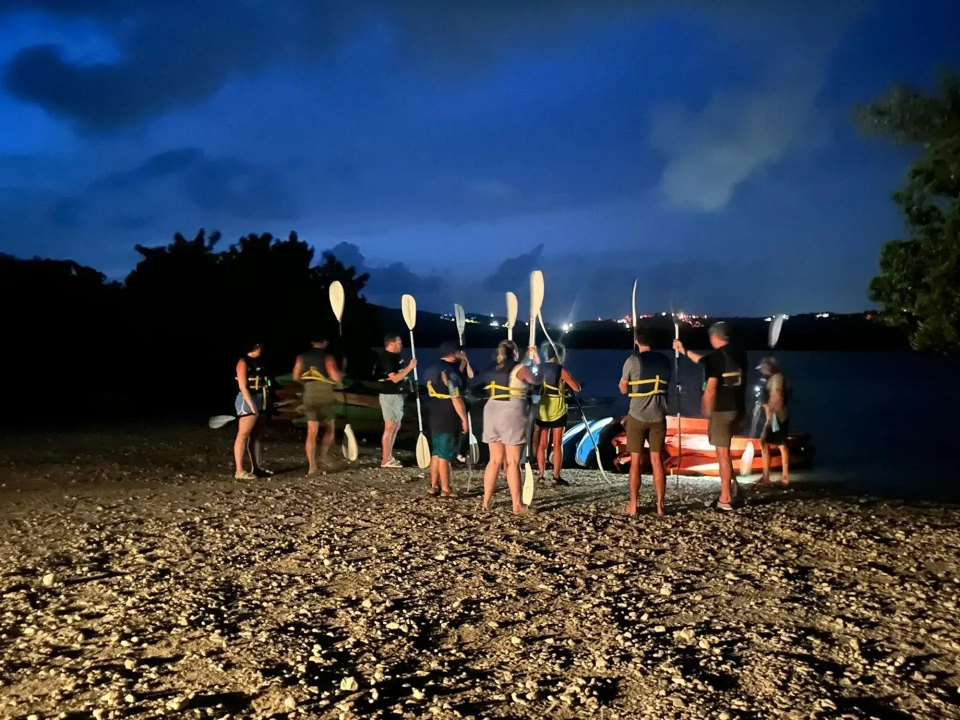 Group of people with paddles gathered on a rocky beach at night beside lit kayaks under a blue sky