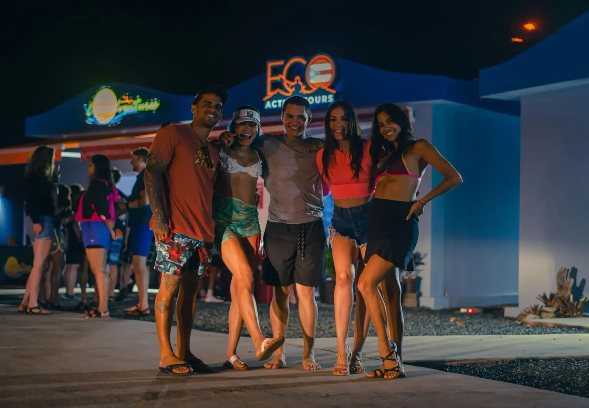 Five people posing outside a neon-lit nightclub at night, with others gathered in the background