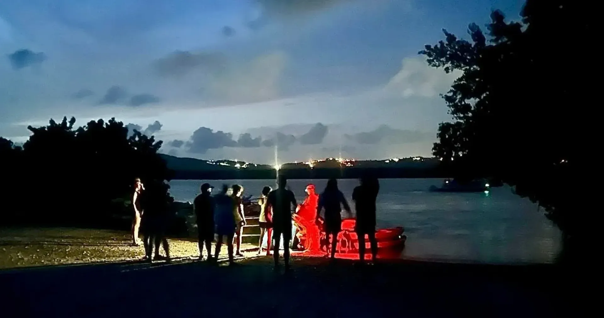 People gathered by a lakeside at dusk, lit by red lights with a glowing horizon in the background