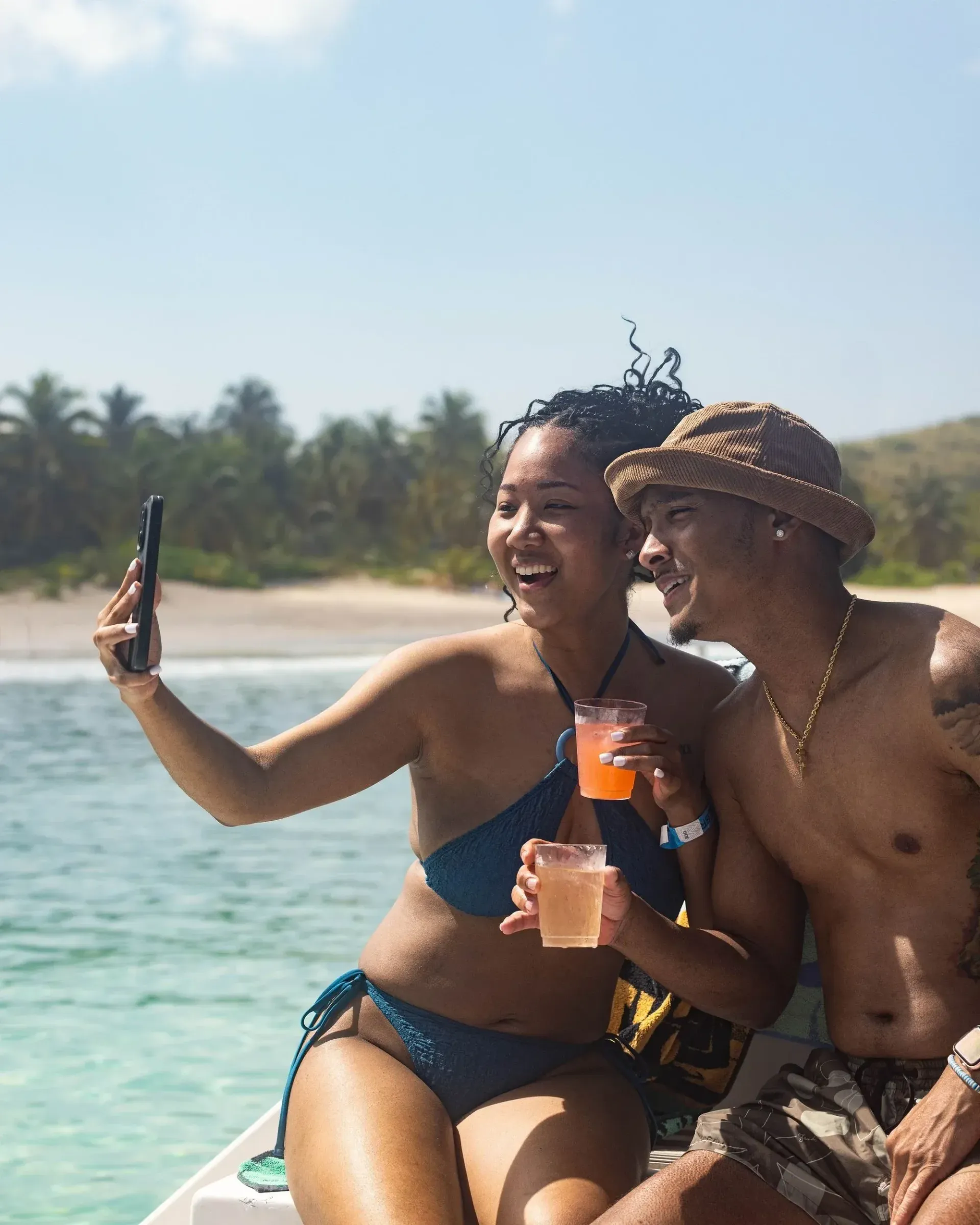 Two people on a boat smiling and taking a selfie with drinks, tropical beach in the background