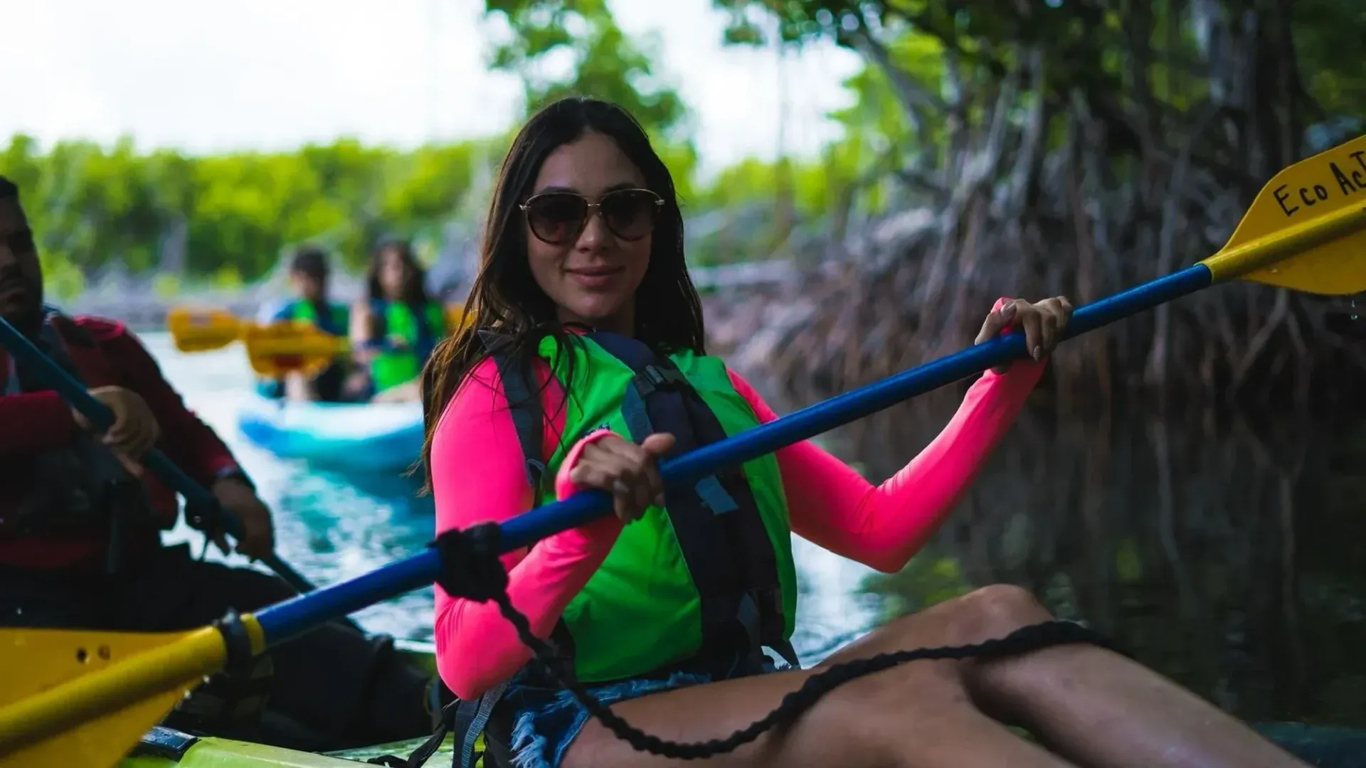 Woman in a kayak wearing a bright pink and green life vest, paddling on a calm river-lined waterway.