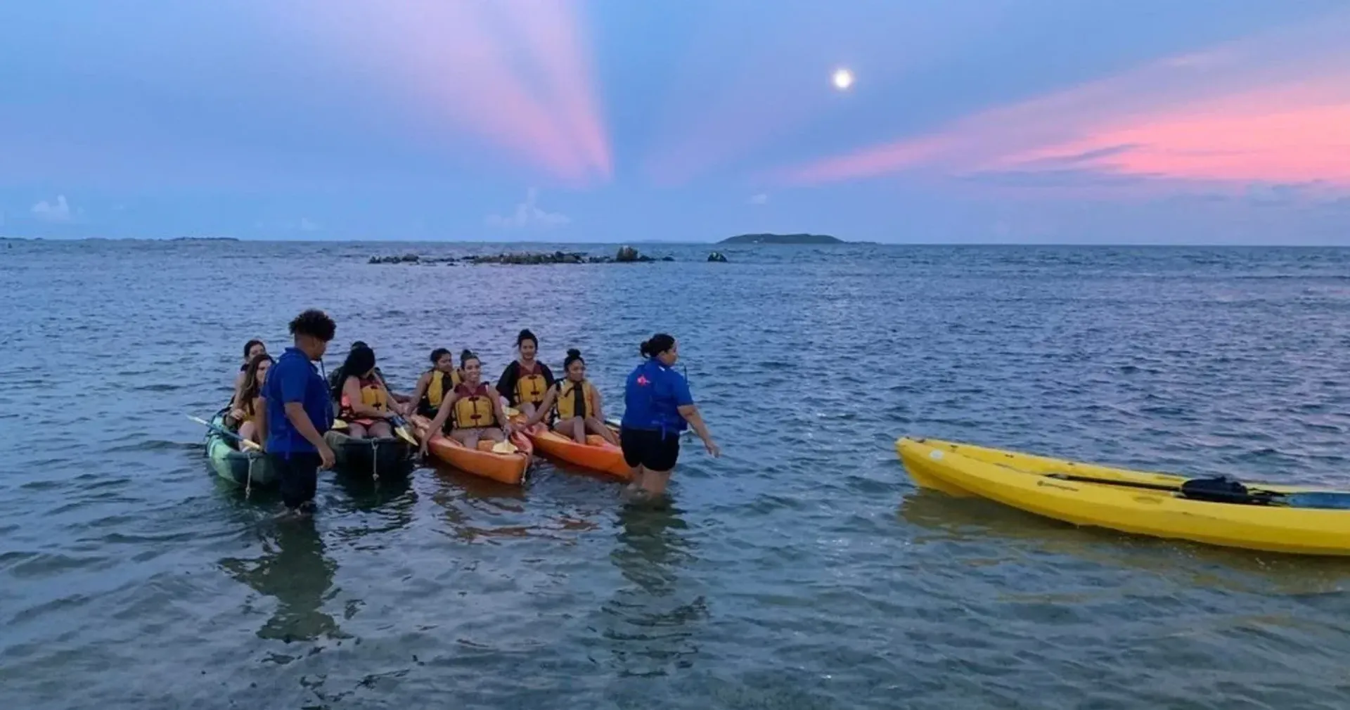 People in life jackets paddle a yellow outrigger boat on calm water at dusk