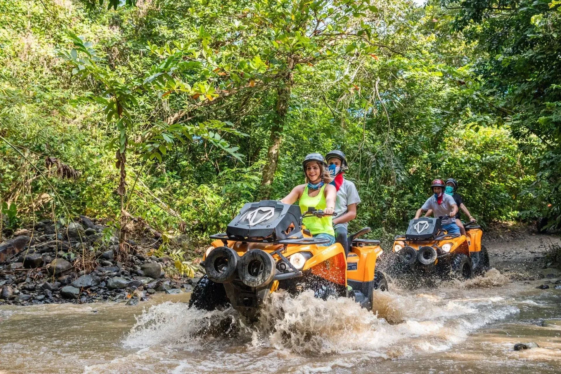 Two orange ATVs driving through a muddy stream in a lush forest with water splashing around them