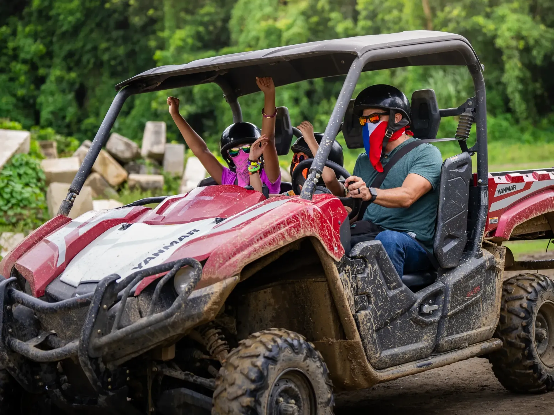 Two people in a red off-road buggy, wearing helmets and face coverings, driving through muddy terrain.