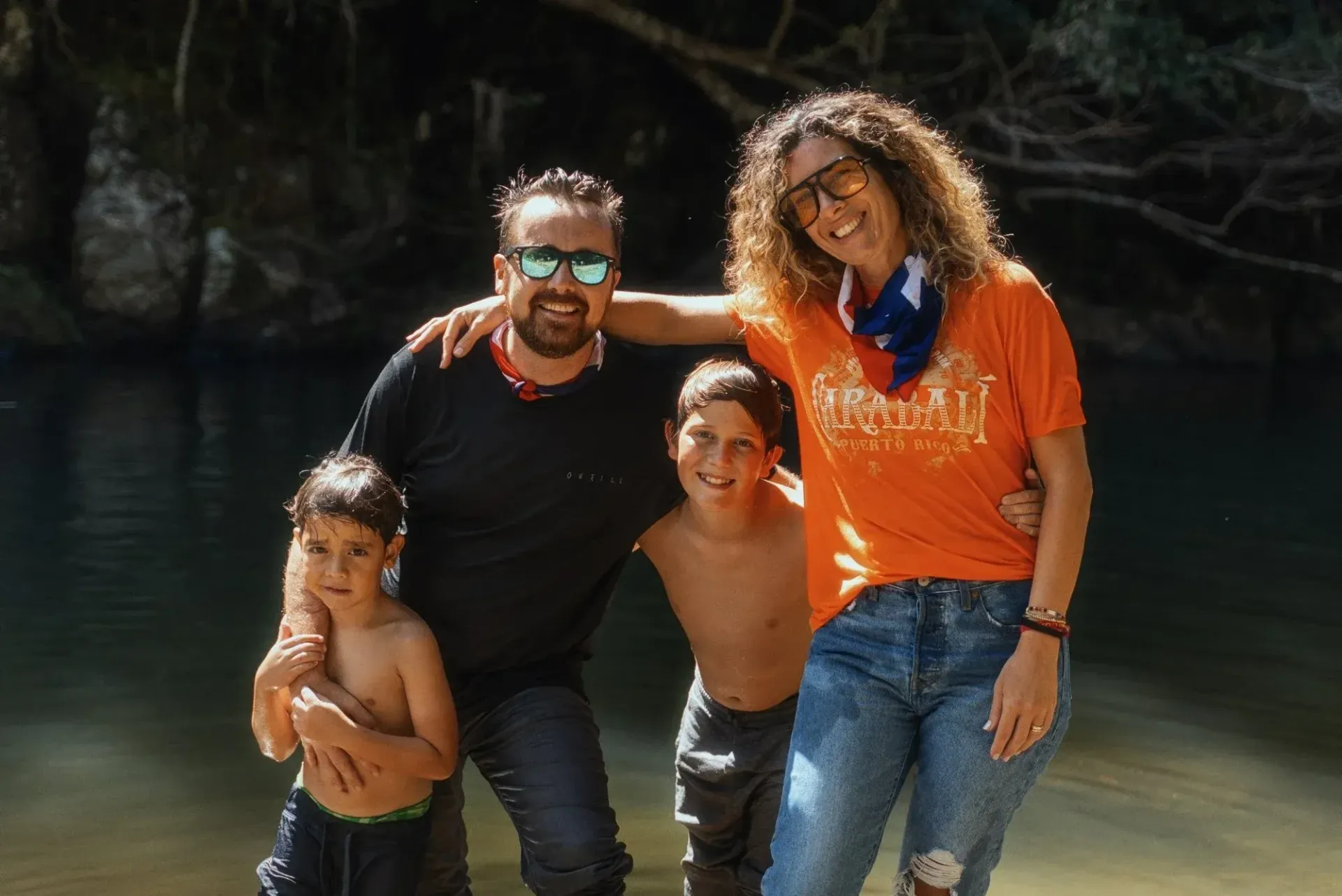Four people posing and smiling outdoors by a river, two in black shirts and two in orange and white.