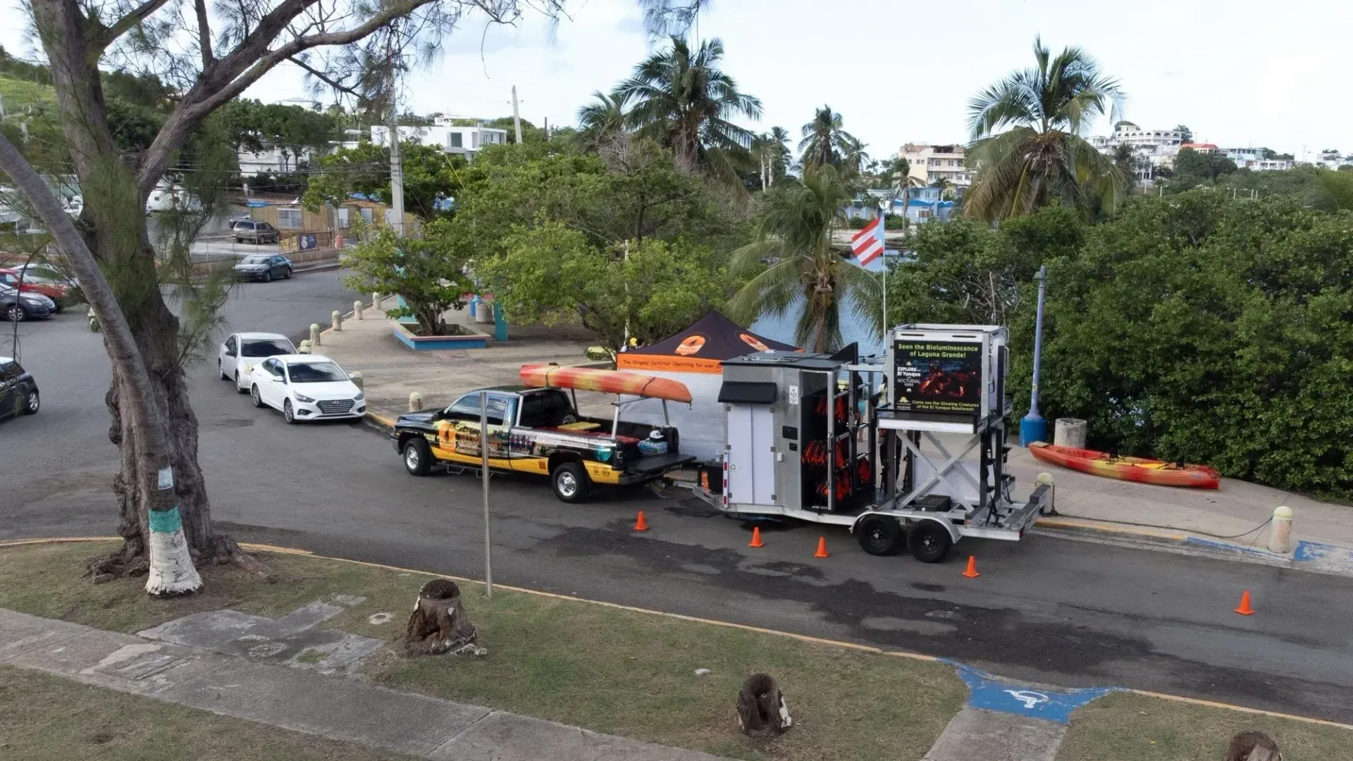 Utility equipment parked on a road beside a sidewalk and trees, with a raised platform truck and cones.