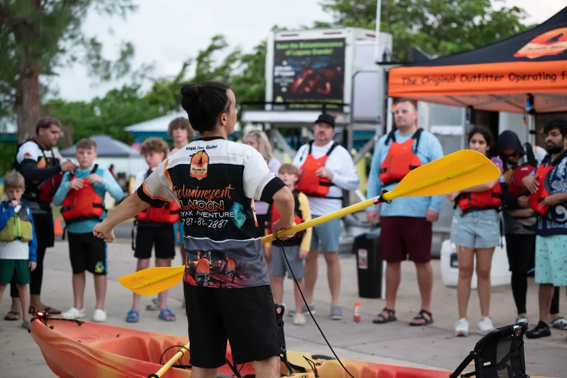 Kayaker in black shirt demonstrates paddle strokes before a small crowd at an outdoor event