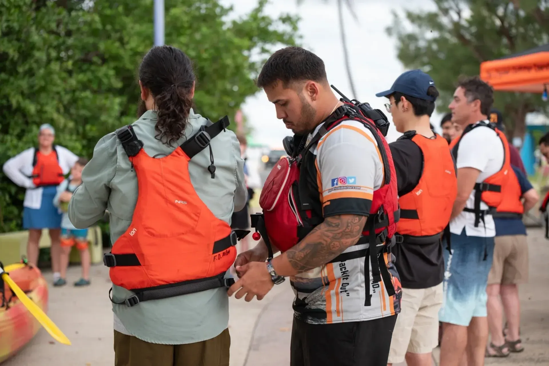 People in orange life vests gathered outdoors, with one person adjusting another’s gear.