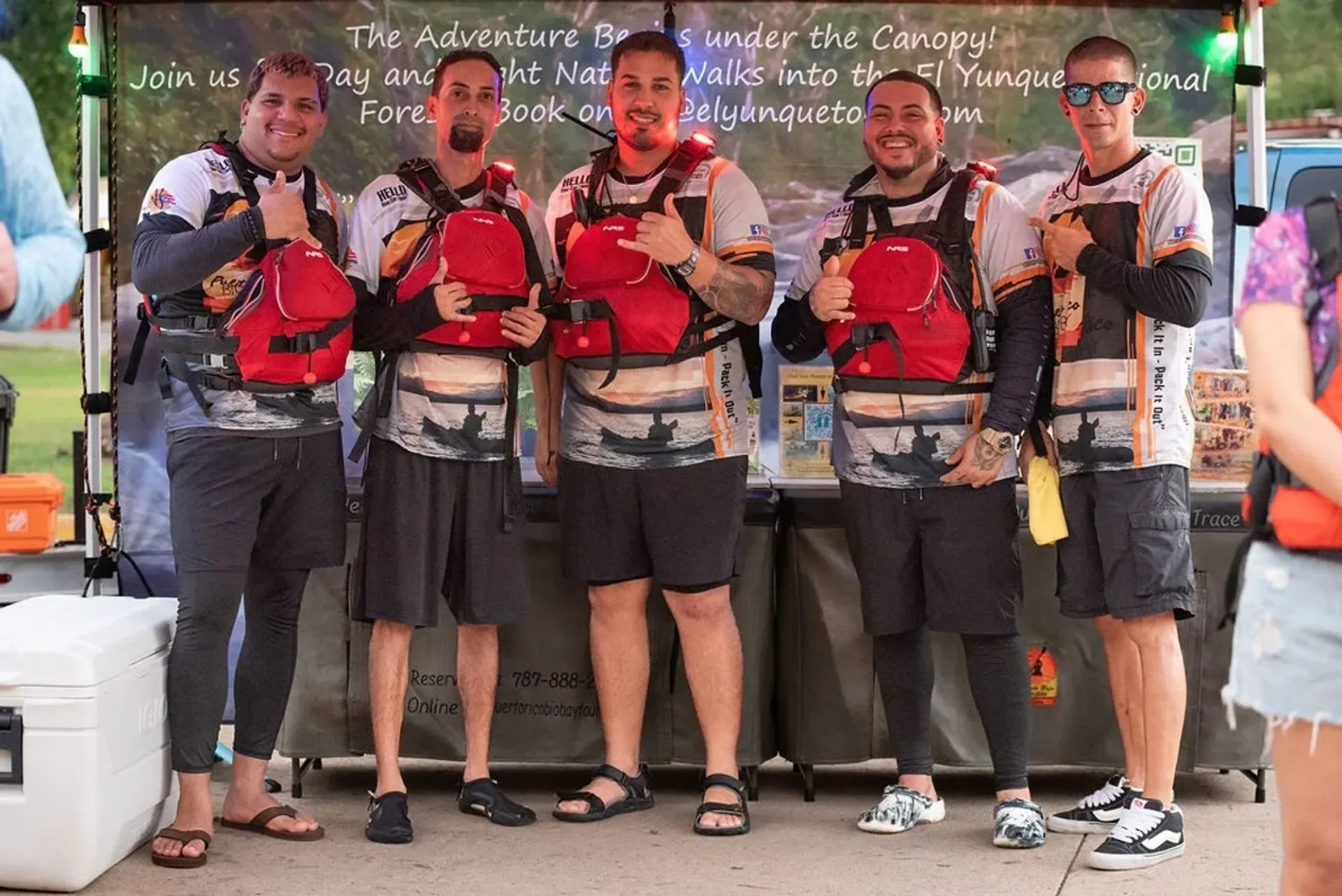 Five people in red life jackets pose with paddleboards in front of a storefront