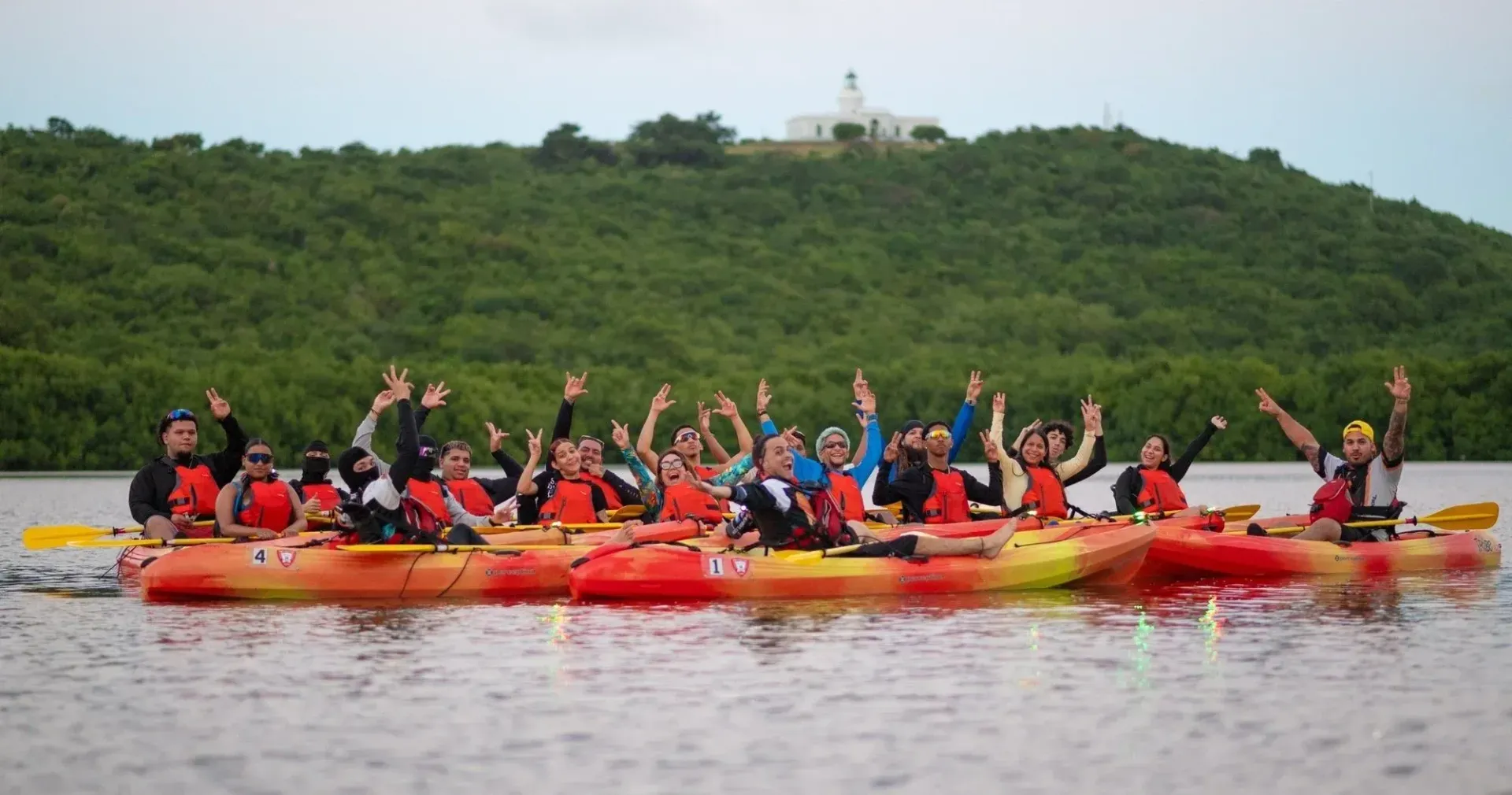People kayaking on a lake in bright red and yellow boats, with a green hill in the background.