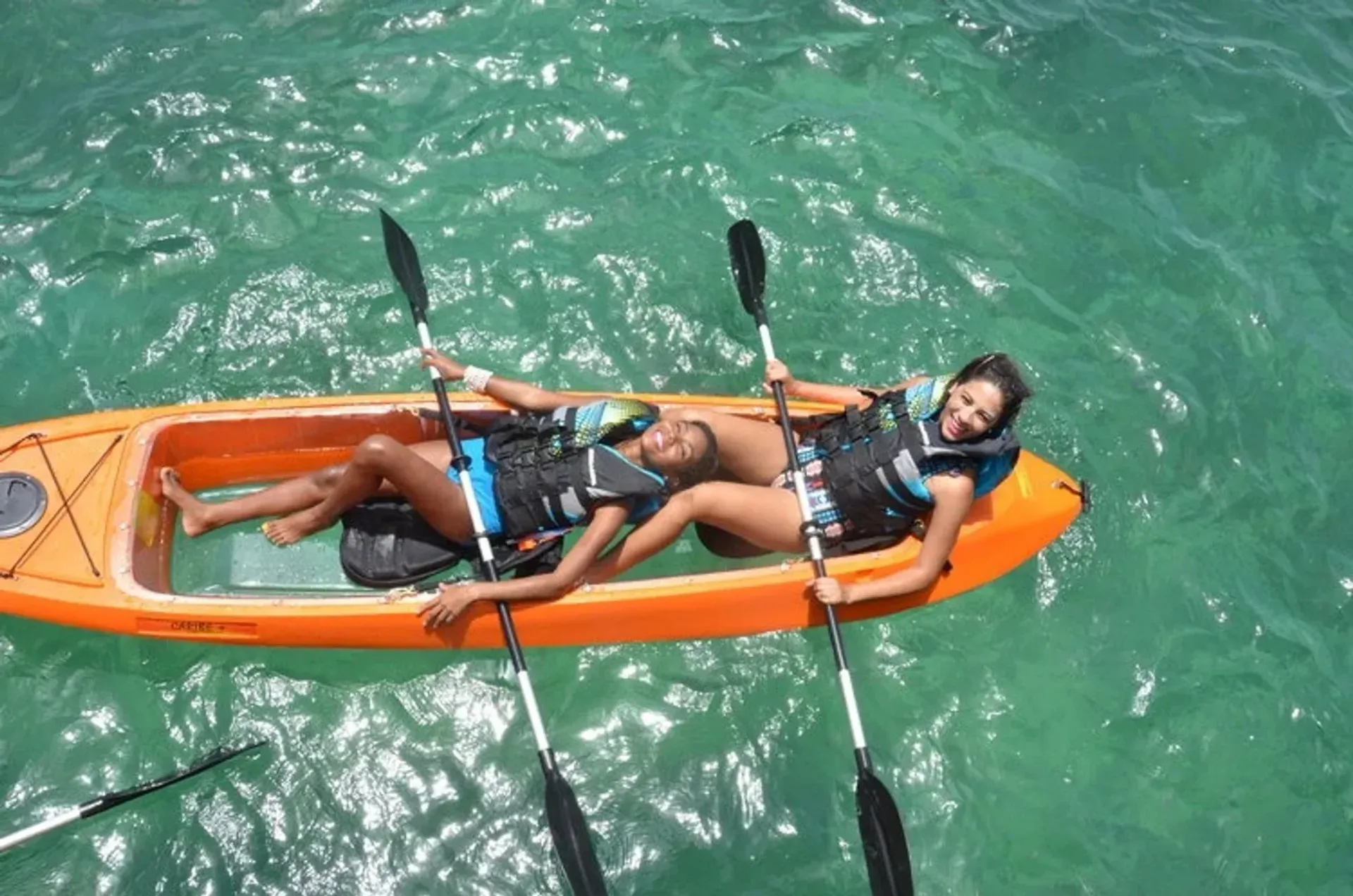 Person kayaking on a calm river beneath overhanging tree branches, with white containers floating nearby.