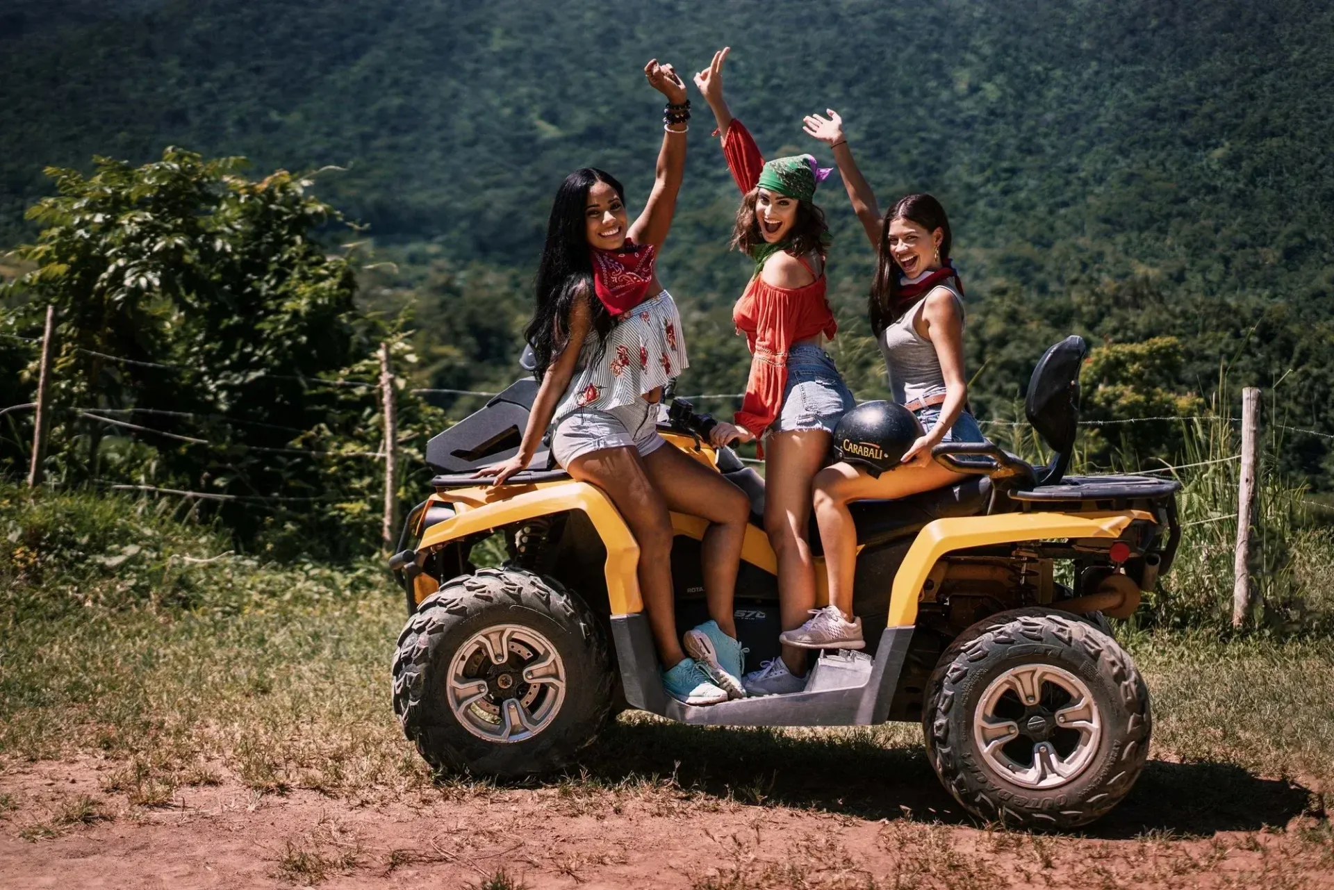 Four friends posing on a yellow ATV on a dirt road, with mountains and trees in the background.