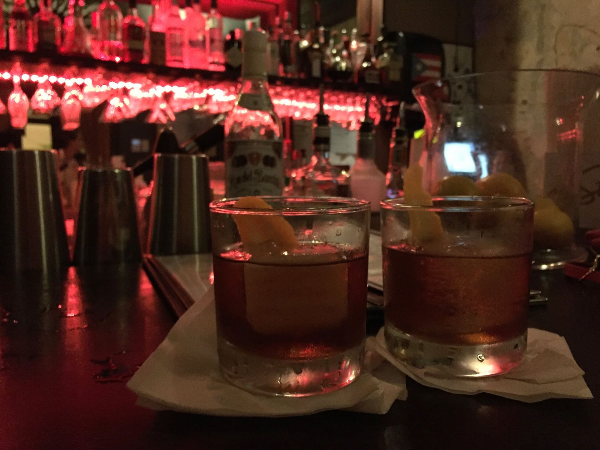 Two cocktails on napkins at a dimly lit bar with red lights and bottles in the background