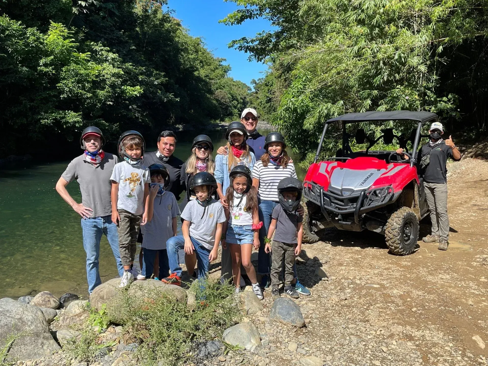 Group posing beside a red off-road vehicle on a muddy trail by a river, with trees in the background