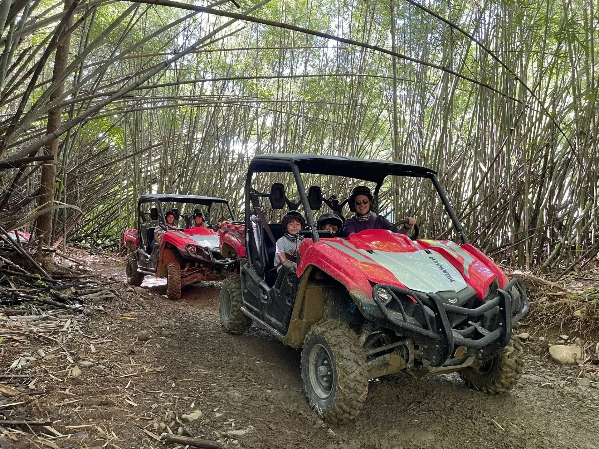 Two red off-road vehicles driving through a wooded trail with people inside