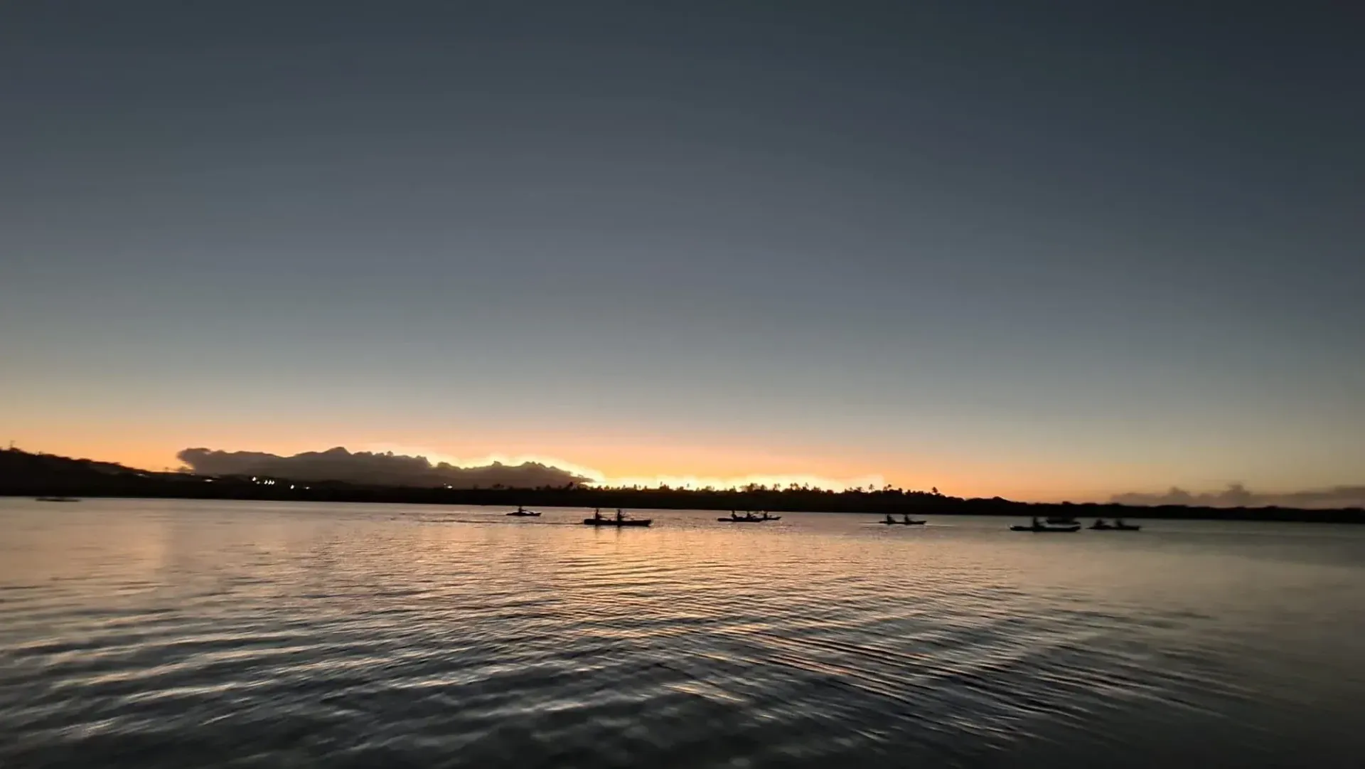Sunrise over a calm lake with silhouetted distant boats and a glowing orange horizon