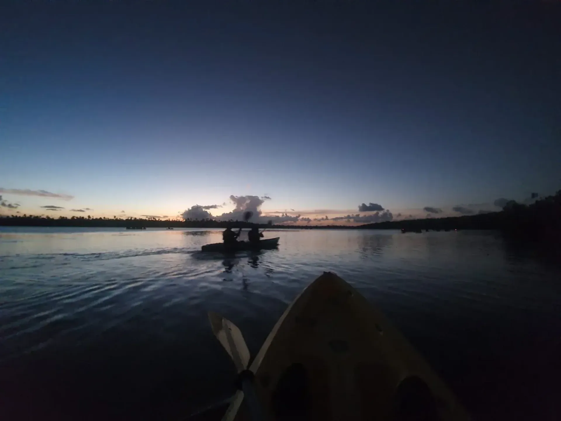 Kayak on calm water at sunset, with a silhouetted dock and clouds on the horizon.