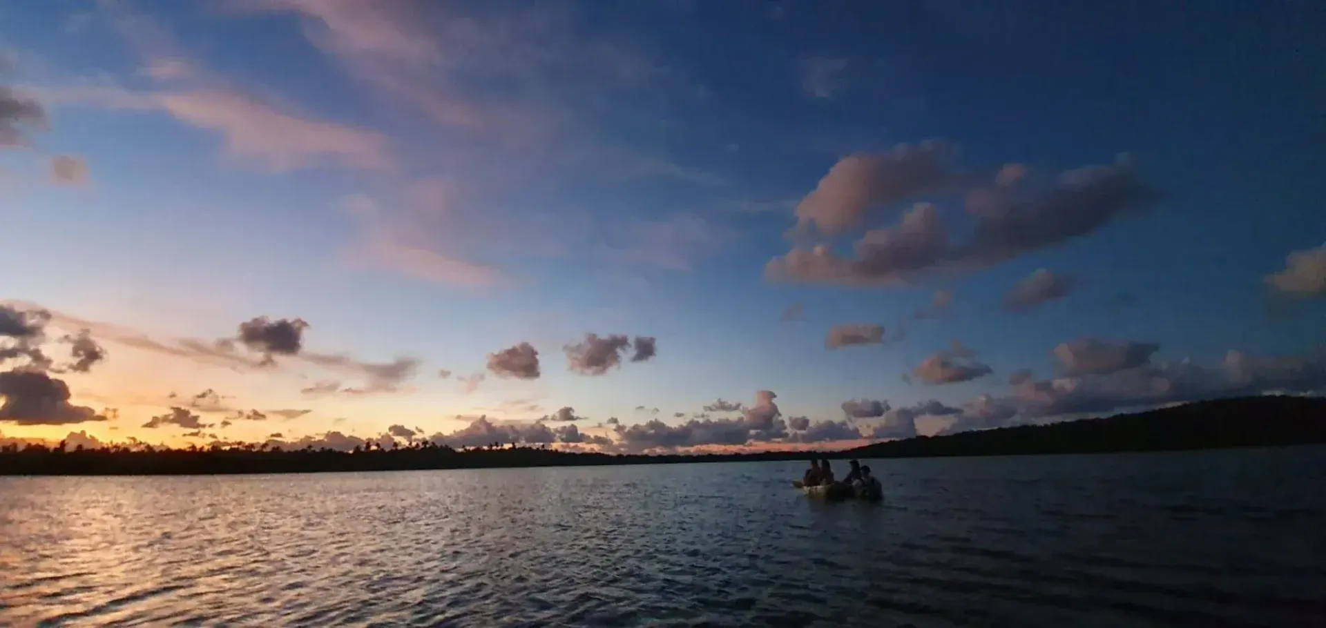 Sunset over calm water with a small boat silhouetted near the horizon and scattered clouds overhead