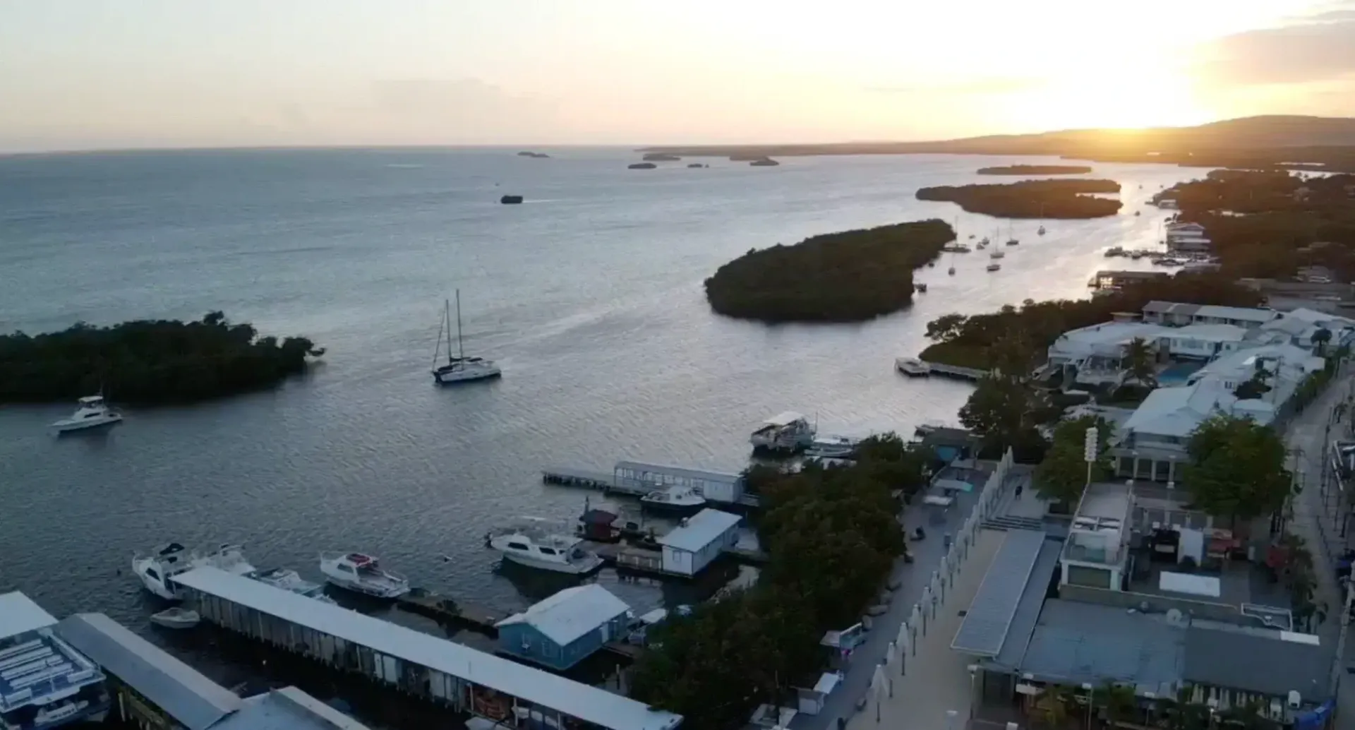 Sunset over a calm bay with boats, docks, and a coastal road lined with buildings