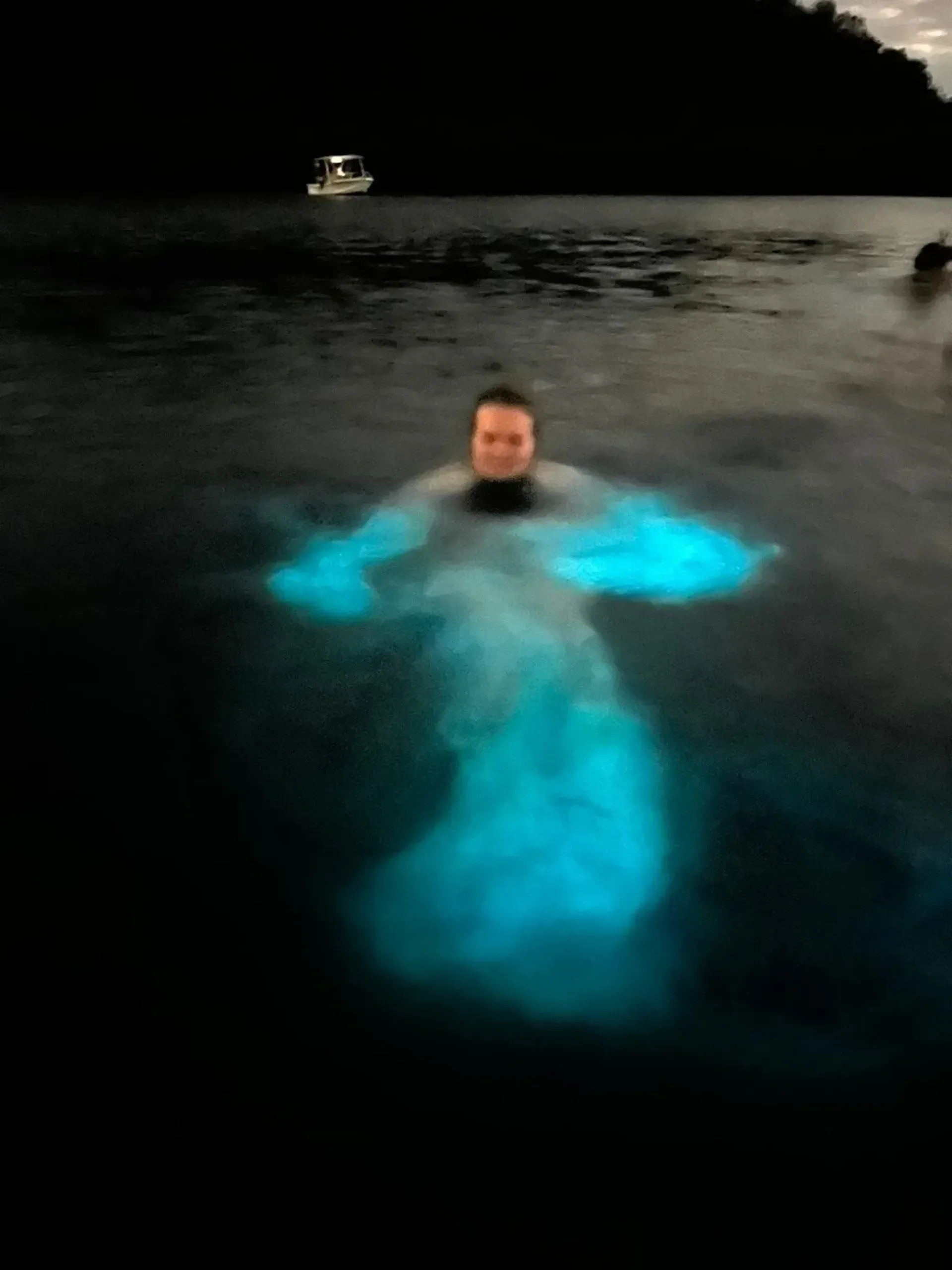 Person swimming at night, glowing turquoise in dark water near a distant boat