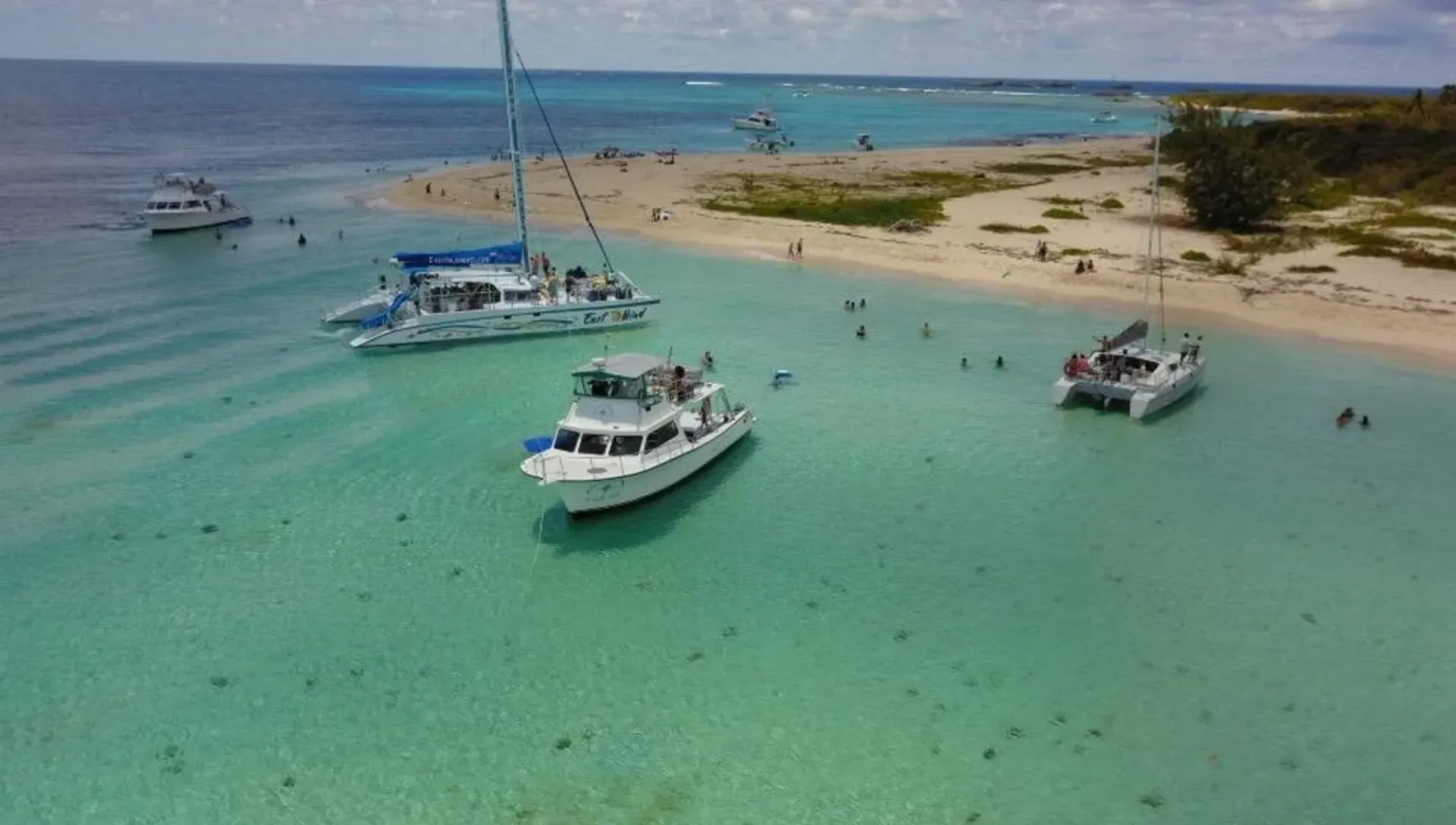 Boats anchored in turquoise water near a sandy beach with people swimming and a sailboat in the background