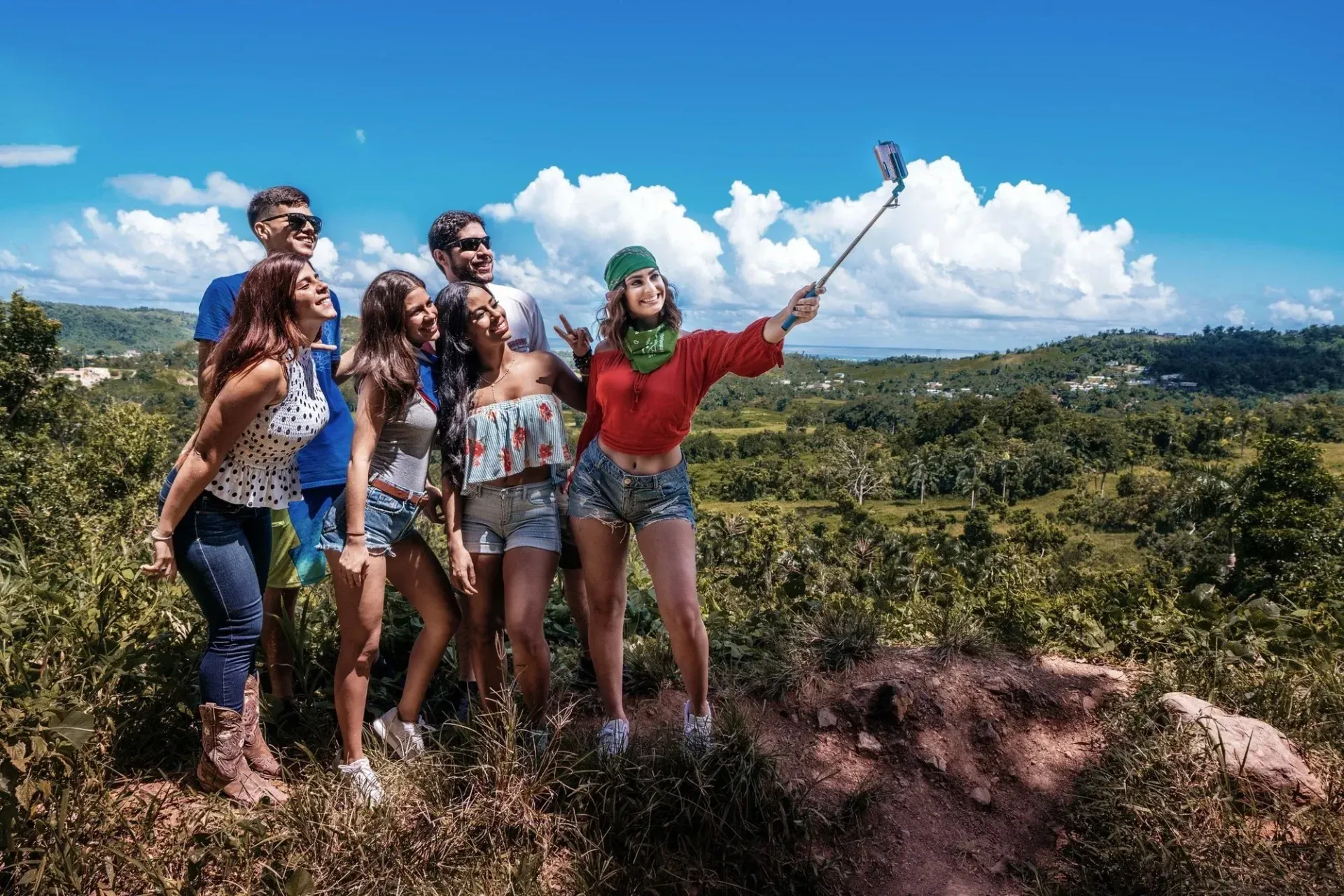 Four people take a selfie on a sunny hilltop overlooking a green landscape and blue sky