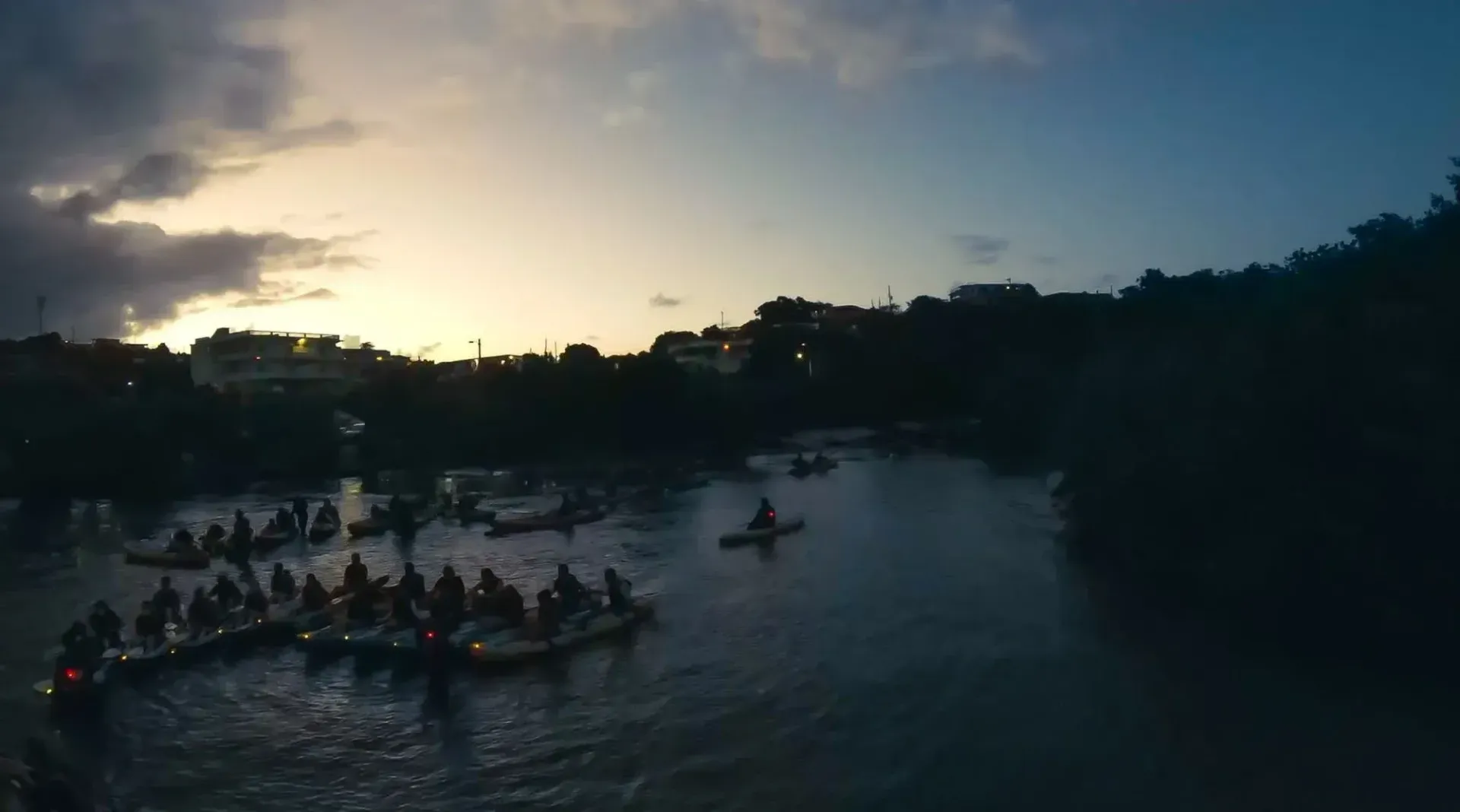 People canoeing on a river at dusk, with a glowing sunset and dark trees along the banks.