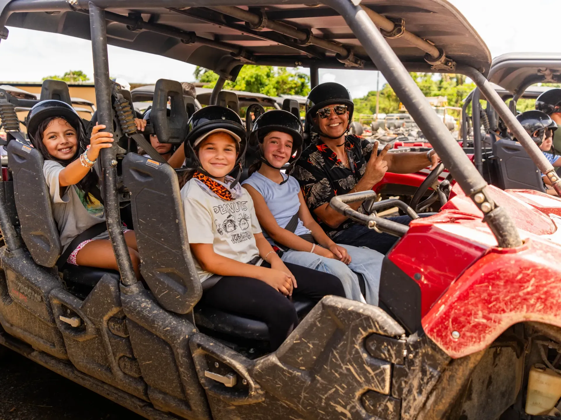 Four people smiling in a red off-road buggy under a canopy, with another vehicle beside them.