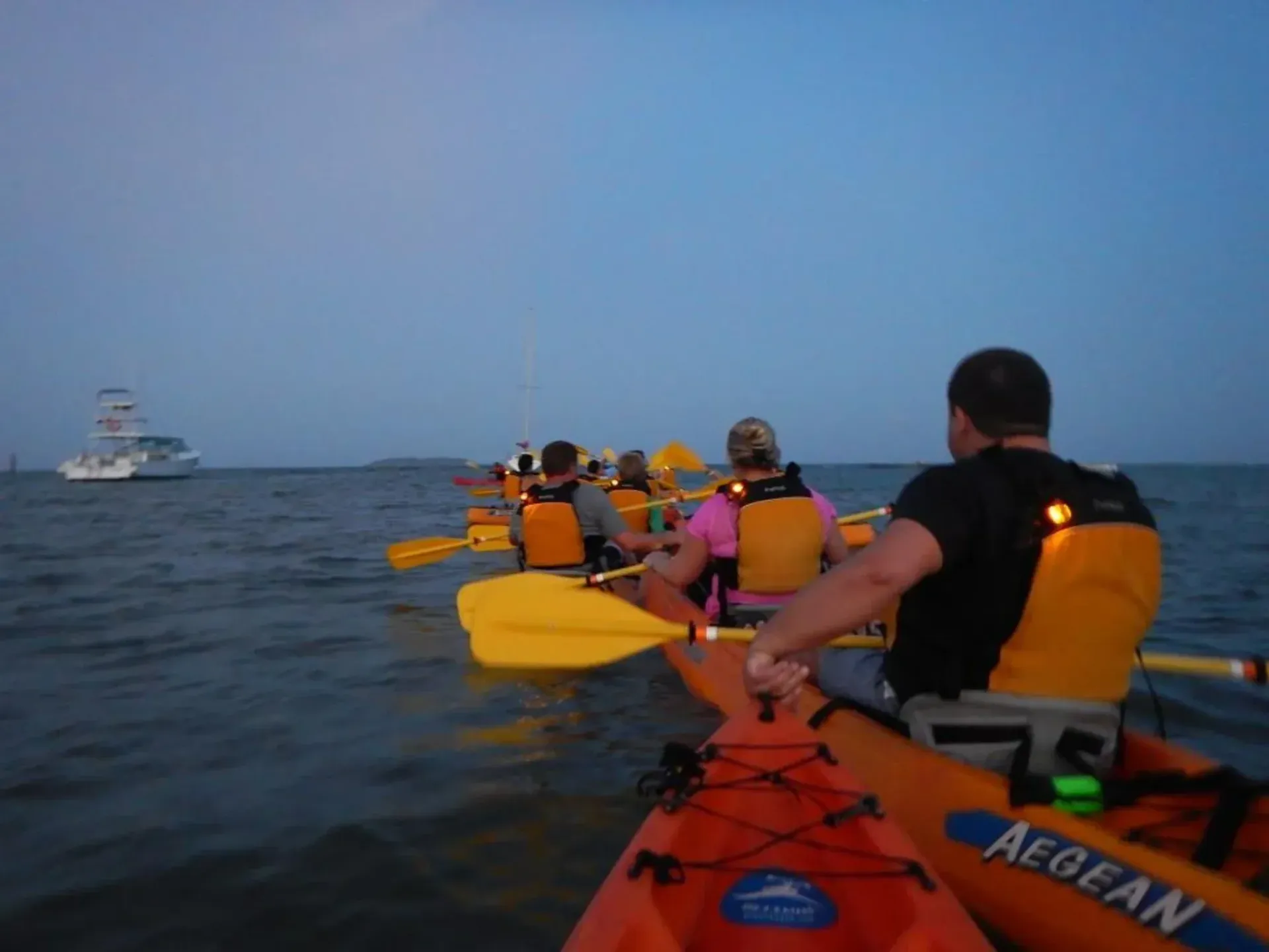 Kayakers in orange life vests paddling on open water at dusk, with a boat in the distance.