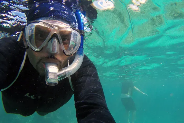 Snorkeler in black wetsuit and mask swimming in clear turquoise water, with another diver in the background