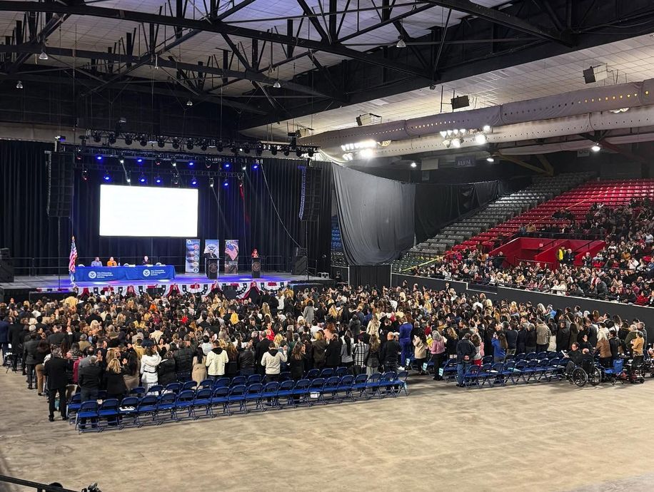 Large crowd at an event with a stage, seating, and a banner. Many people raising their hands.
