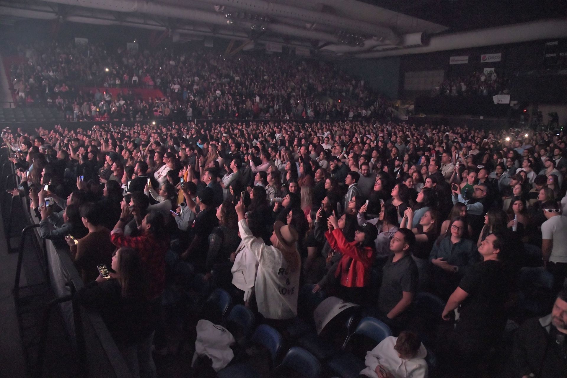 Large crowd at a concert, illuminated by stage lights, with many people raising their hands.