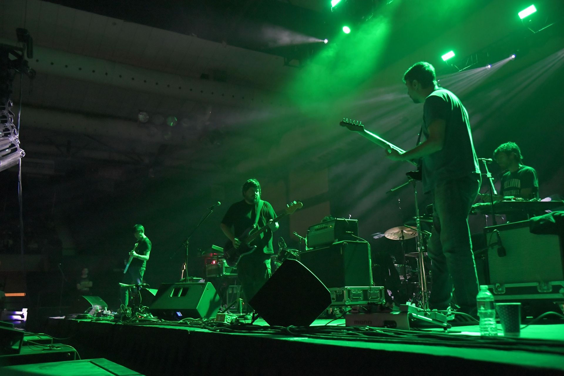 Band performing on a stage with green lighting. Musicians play instruments; drums, guitar, keys, and bass.
