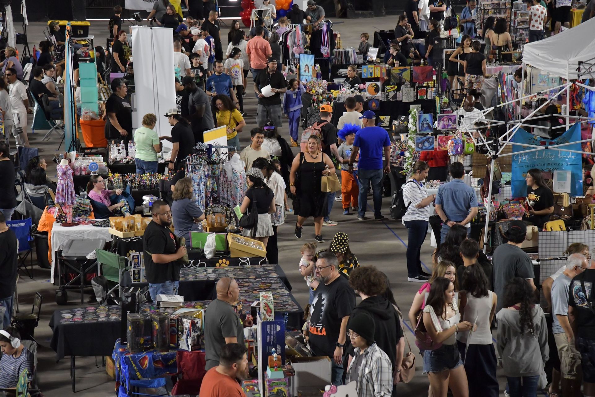 Crowd at an outdoor market, browsing various booths. Goods displayed on tables, people walking around.