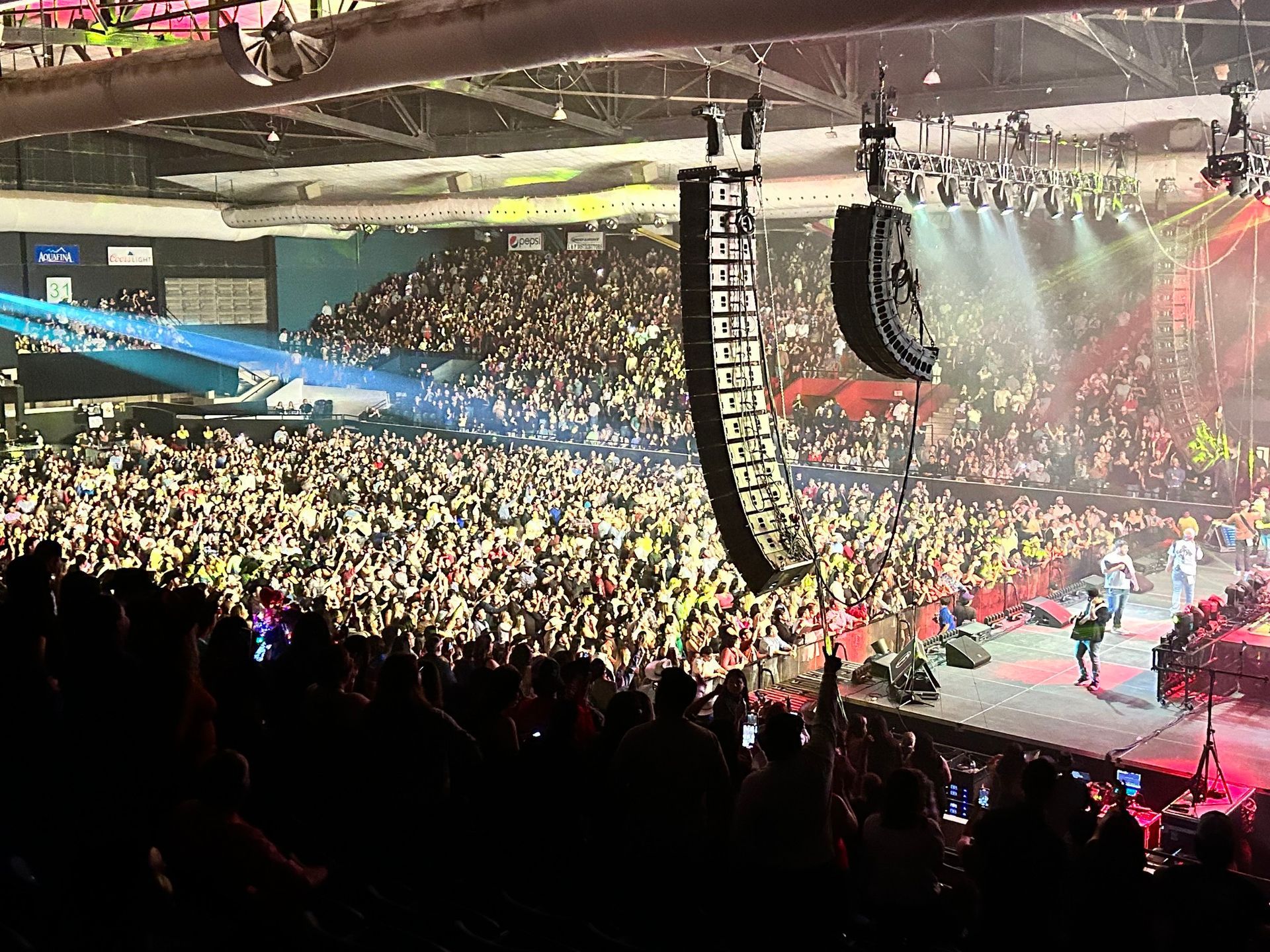 Large crowd at a concert. Stage lit with performers, array of speakers, and an enthusiastic audience.