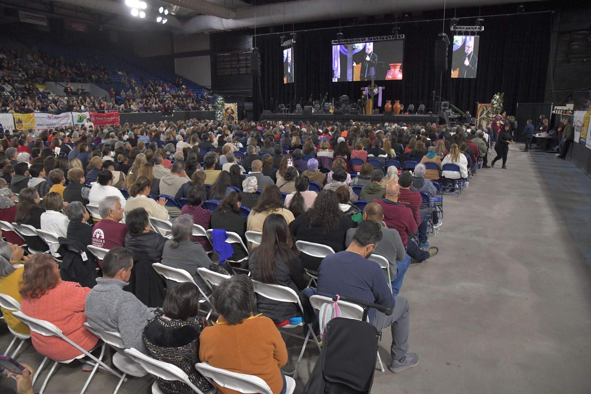 Large audience seated in rows facing a stage. People are listening to speakers and watching screens.