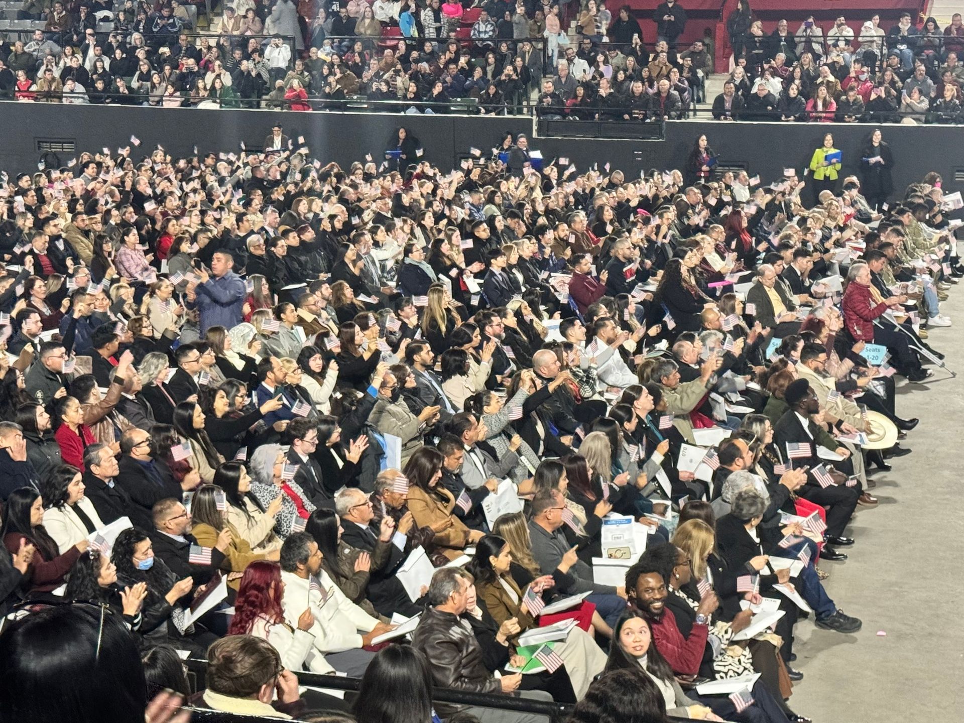 Large audience clapping in an arena; many people, seated, some with papers.
