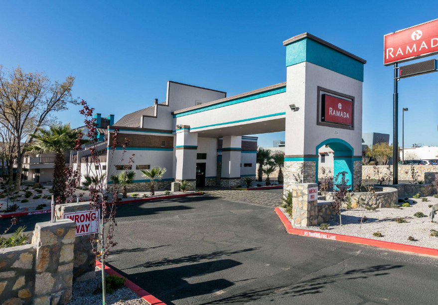 Ramada hotel entrance with teal accents, blue sky, and a parking lot with a sign.