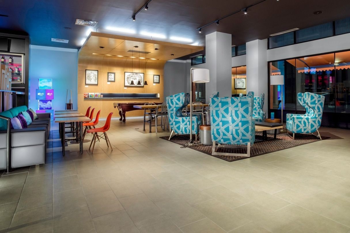 Hotel lobby with seating areas, pool table, and vending machine. Blue and orange accents, neutral tones.