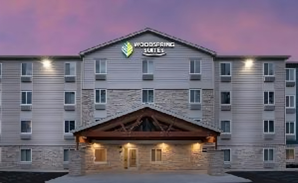 WoodSpring Suites hotel exterior at dusk. Gray facade, lit entryway, logo above entrance.