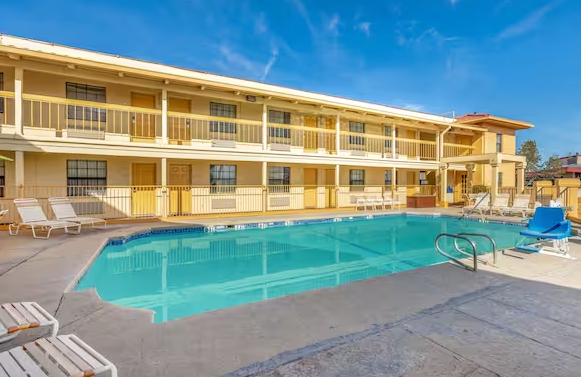 Motel with pool; two-story building with balconies, beige exterior, turquoise water. Chairs line the pool deck.