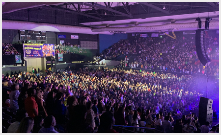 Large crowd at an indoor concert, purple lighting, hands raised, watching the stage.