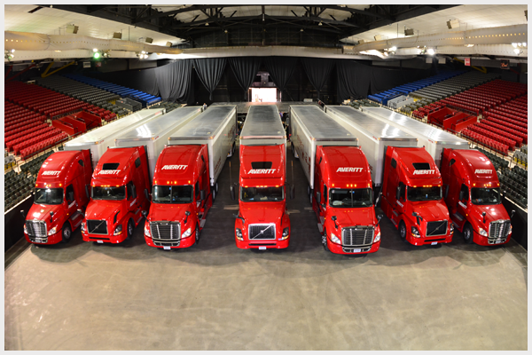 Red semi-trucks lined up on a floor inside a stadium, with red and blue seating.