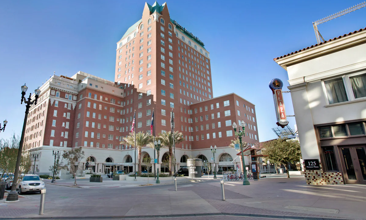Large red brick hotel, palm trees, and a plaza under a clear blue sky.