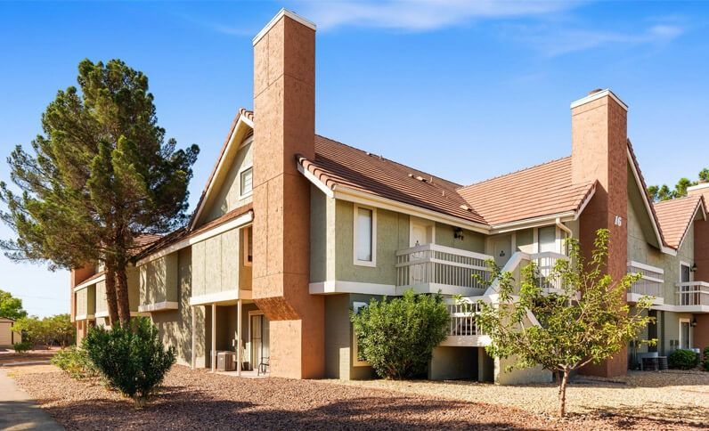 Two-story townhouses with red tile roofs, light green siding, and tall brick chimneys, on a gravel lot, sunny day.