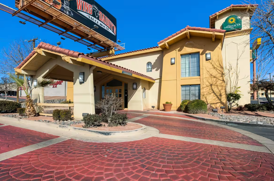 Exterior of a beige hotel with a red brick driveway, under a billboard, blue sky.