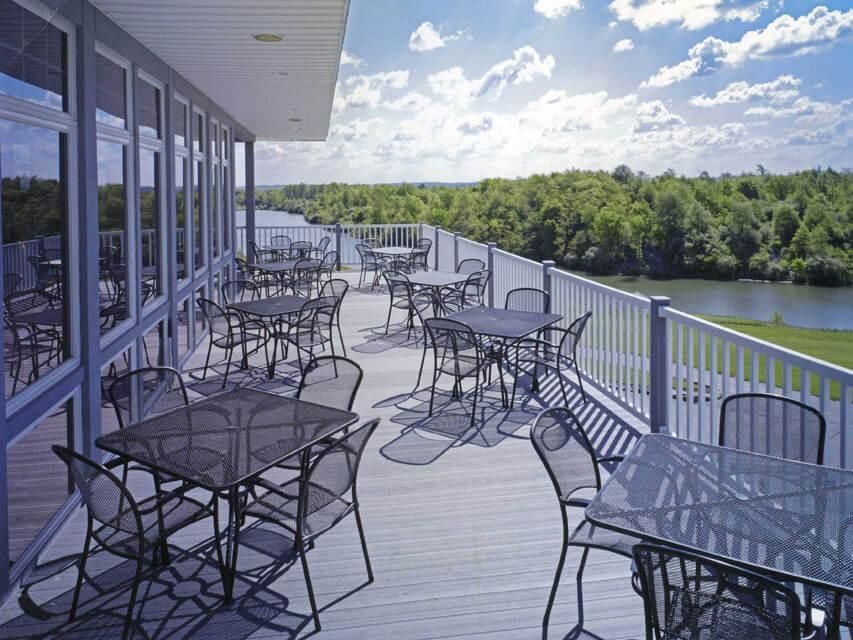 Outdoor restaurant patio with tables and chairs overlooking a river and trees under a sunny sky.